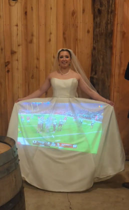 Com paredes de madeira escura no local do casamento, Kennedie teve uma ideia inusitada. Ela transformou seu vestido de noiva em uma tela para projetar o jogo durante a recepção (Foto: Reprodução/TikTok/@lost_in_the_trees_media)