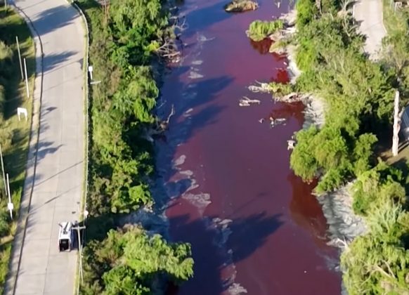 A proximidade de um depósito industrial aumentou as suspeitas sobre descarte irregular de resíduos. Autoridades locais iniciaram uma investigação para esclarecer o caso. Amostras da água foram coletadas para análise e determinar a origem da substância que tingiu o rio (Foto: Reprodução/CNN)